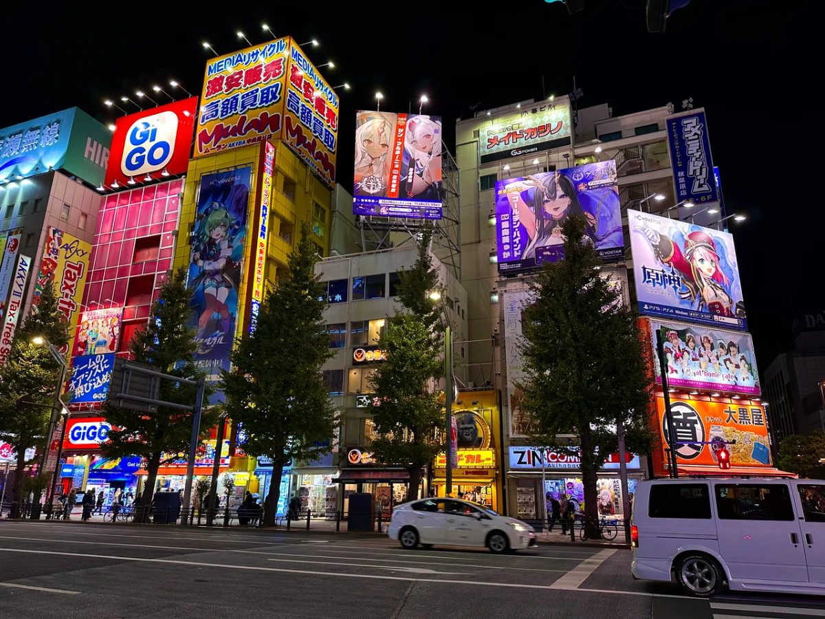 Night view of a busy Tokyo street lined with brightly lit buildings displaying colorful anime and game advertisements.