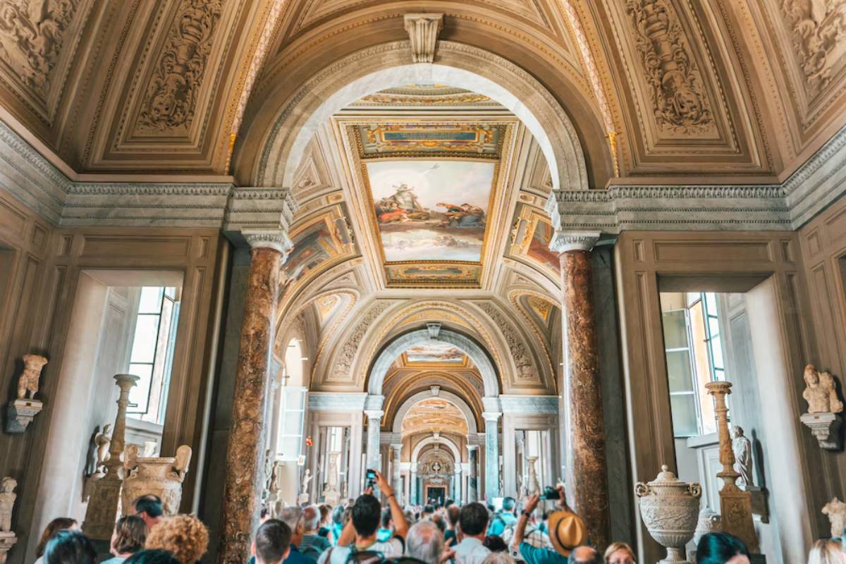 Tourists inside a museum with ornate ceilings and classical sculptures lining a grand hallway.