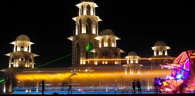 Illuminated structures and a glowing art car at a night festival, with people and colorful laser beams.