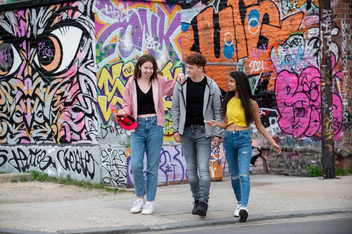 Three young adults walking and smiling together in front of a colorful graffiti wall.