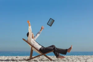 Man in a suit joyfully tossing a tablet into the air while relaxing on a beach chair with a clear blue sky above.