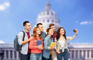 Group of six young adults taking a selfie in front of the U.S. Capitol building, smiling and holding books and backpacks.