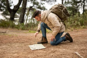 A man with a backpack kneels in the woods using a compass on a map.