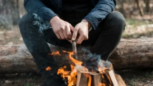 Man in dark clothing sits by a small campfire, poking it with a stick, surrounded by woods.