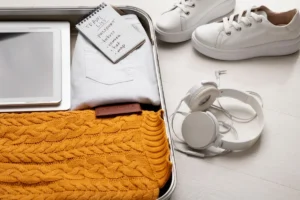 Open suitcase with clothes, white shoes, headphones, tablet, and travel list on a wooden floor.