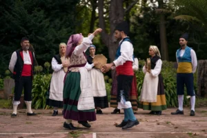 People in traditional European costumes dance outdoors, watched by others playing tambourines and observing.
