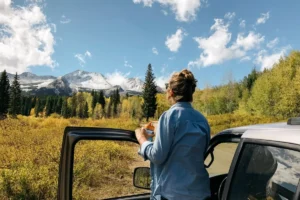 Person standing by a car, sketching a mountainous landscape with snowy peaks and autumn trees.