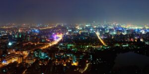 Nighttime aerial view of a vibrant, brightly lit cityscape with dense buildings and a long illuminated road.