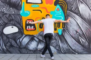 Man with arms extended in front of a colorful, abstract street mural.
