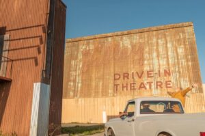 A person enthusiastically reaching out of a white truck at a rustic "Valley Drive-In Theatre."