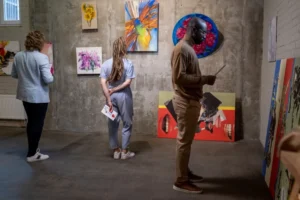 Three people viewing various colourful paintings at an art exhibition in a gallery with concrete walls.