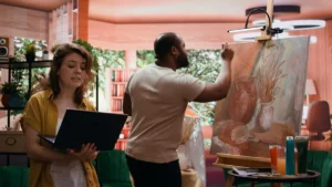A man painting on an easel in a colourful studio, with a woman holding a laptop observing his work.
