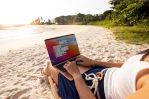 Person using a laptop on a sunny beach, sitting on a wooden chair with sandy beach and greenery in the background.