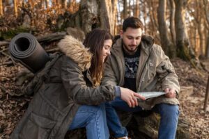 Two people sitting in a forest, looking at a map together, dressed in casual winter wear.
