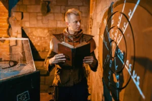 A man in medieval attire reads a book beside a dartboard with glowing runes in a dimly lit room.