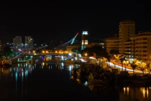 Night view of a city riverfront with illuminated buildings, a bridge with light streaks, and moored boats reflecting in the water.